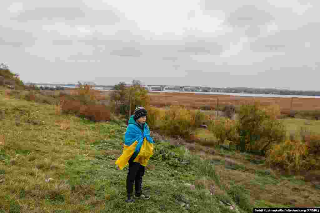Хлопчик, з українським прапором на спині, стоїть неподалік зруйнованого Антонівського мосту в перші дні звільнення Херсона від російських військ. Листопад 2022 року